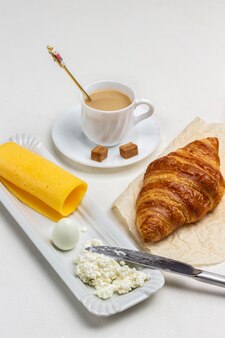 Rogalik i filiżanka kawy. twarożek i nóż, ser i jajko. produkty śniadaniowe. białe tło. widok z góry