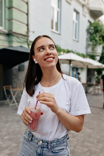 Romantyczna atrakcyjna kobieta ze szczęśliwym szerokim uśmiechem trzymająca koktajl i patrząca w górę z wielkimi emocjami na świeżym powietrzu w letnim mieście