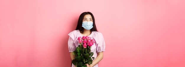 Romantyczna azjatycka dziewczyna w masce medycznej patrząca na pustą przestrzeń z marzycielskimi oczami trzymająca bukiet