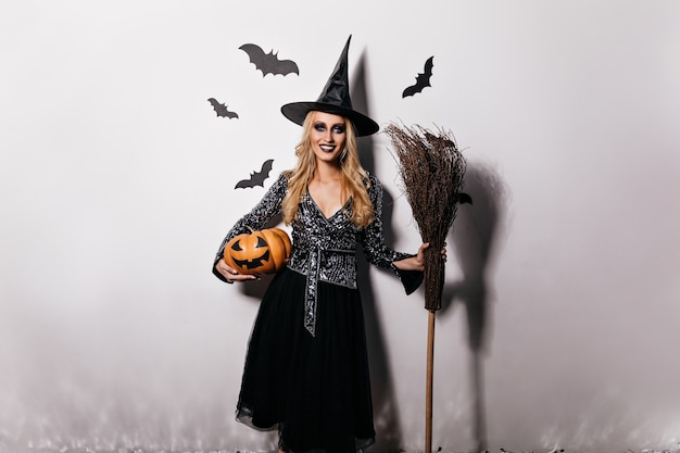 Romantyczna Blondynka Pozuje Z Dynią I Nietoperzami. Kryty Strzał Uśmiechnięta Młoda Kobieta Korzystających Z Karnawału Halloween.