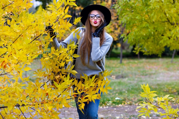 Romantyczna długowłosa dziewczyna pozuje z wyrazem twarzy pocałunek podczas spaceru w parku jesienią. Zewnątrz portret eleganckiej europejskiej młodej kobiety w dżinsach i kapeluszu stojącym obok żółtego krzewu.