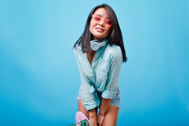 Romantyczna dziewczyna azjatyckich z lśniącymi prostymi włosami pozuje z przyjemnością. Ładna modelka europejska w okulary przeciwsłoneczne i słuchawki, uśmiechając się, spędzając czas.