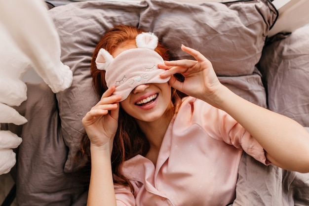 Bezpłatne zdjęcie romantyczna dziewczyna leży w łóżku z natchnionym uśmiechem ujęcie z góry wesołej rudej damy.