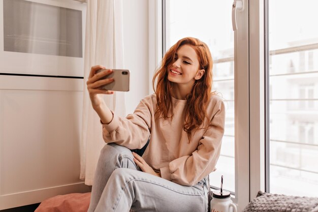 Romantyczna dziewczyna w dżinsach robi selfie w domu Zwinna ruda młoda dama robi sobie zdjęcie