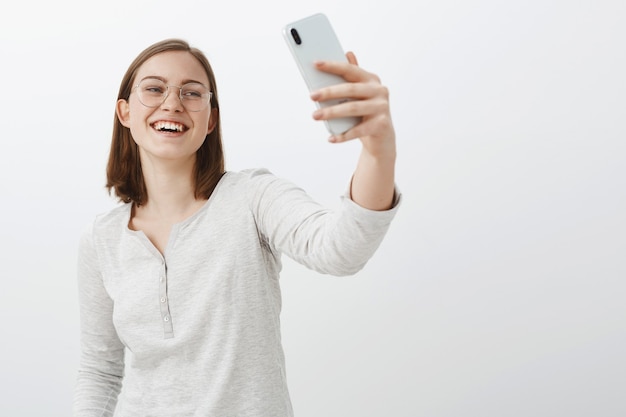 Rozbawiona, dobrze wyglądająca europejska studentka z krótkimi brązowymi włosami w przezroczystych okularach, trzymająca smartfona i śmiejąca się, patrząc na ekran urządzenia rozmawiająca przez wiadomości wideo na szarej ścianie