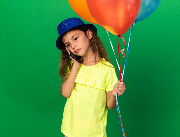 rozczarowana mała dziewczynka kaukaski z niebieskim kapeluszem strony, trzymając balony z helem i rozmawiając przez telefon na białym tle na zielonej ścianie z miejsca na kopię