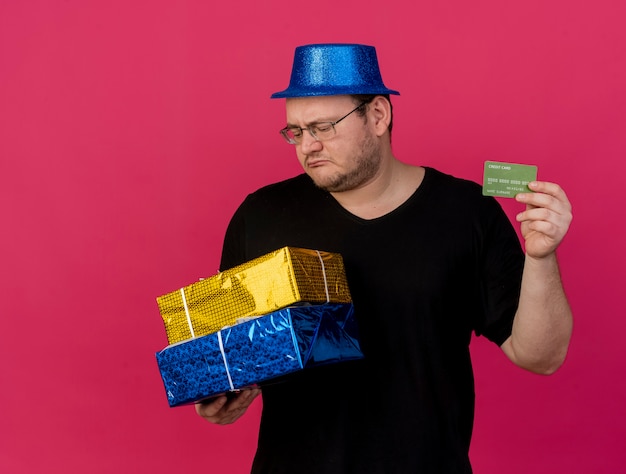 Rozczarowany dorosły słowiański mężczyzna w okularach optycznych w niebieskim kapeluszu imprezowym, trzyma pudełka na prezenty i kartę kredytową