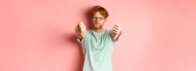 Bezpłatne zdjęcie rozczarowany młody człowiek w okularach z niechlujną czerwoną fryzurą pokazujący kciuki w dół i krzywiący się niezadowolony