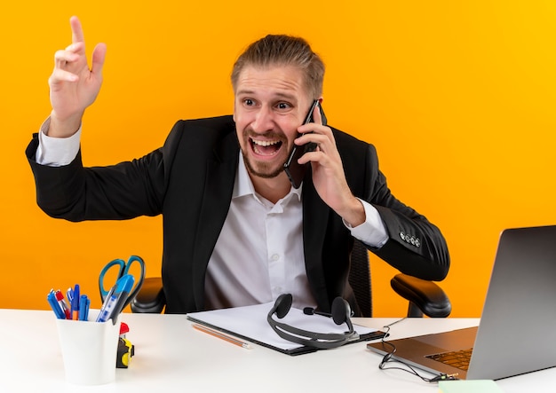 Rozczarowany przystojny biznesmen w garniturze pracuje na laptopie rozmawia przez telefon komórkowy wygląda na zdezorientowanego i niezadowolonego siedzącego przy stole w biurze na pomarańczowym tle