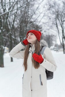 Rozmyślająca młoda kobieta w czerwonej czapce z dzianiny i rękawiczkach spaceruje po zaśnieżonym parku. zimowy dzień.