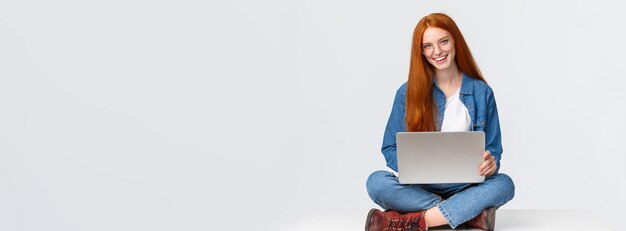 Rozochocona bezczelna ruda studentka college'u dziewczyna w okularach siedzi na skrzyżowanych nogach z laptopem wo