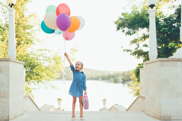 Rozochocona dziewczyna trzyma kolorowych balony i dziecięcą walizkę