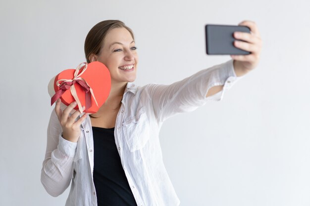 Rozochocona kobieta bierze selfie fotografię z sercem kształtował prezenta pudełko
