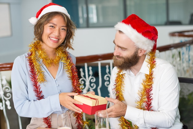 Rozochocony biurowy asystent w Santa kapeluszu daje Bożenarodzeniowemu prezentowi