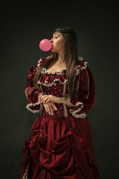 Różowa guma balonowa. Portret średniowiecznej młodej kobiety w czerwonej odzieży vintage stojącej na ciemnym tle. Modelka jako księżna, osoba królewska. Pojęcie porównania epok, nowoczesności, mody, piękna.