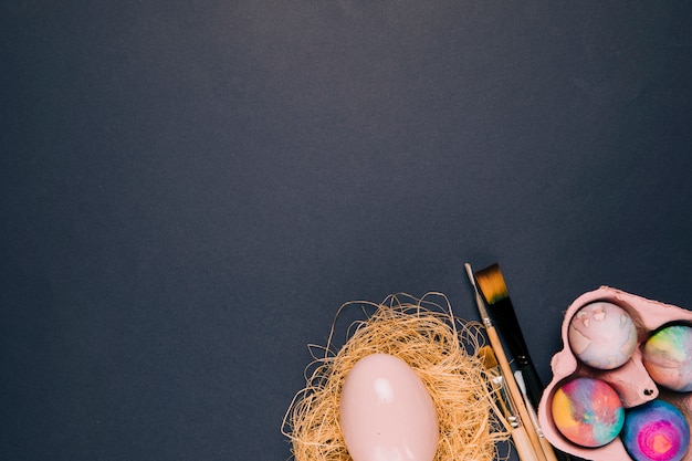 Bezpłatne zdjęcie różowe jajko wielkanocne w gnieździe; pędzle i karton jaj na rogu czarnym tle