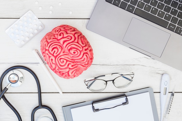 Różowy model mózgu; pigułki; stetoskop; długopis; okulary; schowek; termometr; strzykawka i laptop na drewniane biurko