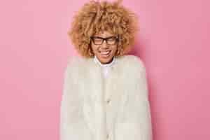 Bezpłatne zdjęcie rozradowana kobieta o blond kręconych włosach czuje się bardzo szczęśliwa, chichocze, pozytywnie nosi przezroczyste okulary, a zimowy płaszcz wyraża szczęśliwe emocje odizolowane na różowym tle. koncepcja szczęścia