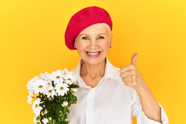 Rozradowana, radosna kobieta w średnim wieku, ubrana w czerwoną czapeczkę z boku, pokazująca kciuki do góry, wyrażająca aprobatę, zachęcająca do kupowania kwiatów.