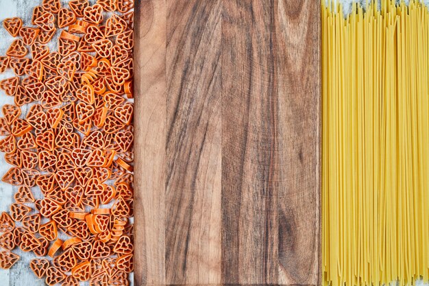 Rozrzucony makaron w kształcie serca i spaghetti wokół drewnianej deski.