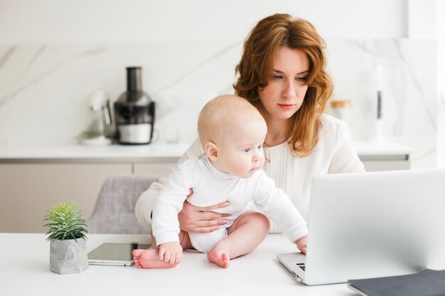 Rozważna biznesowa kobieta siedzi przy stole i pracuje na laptopie, trzymając w pobliżu swoje słodkie małe dziecko