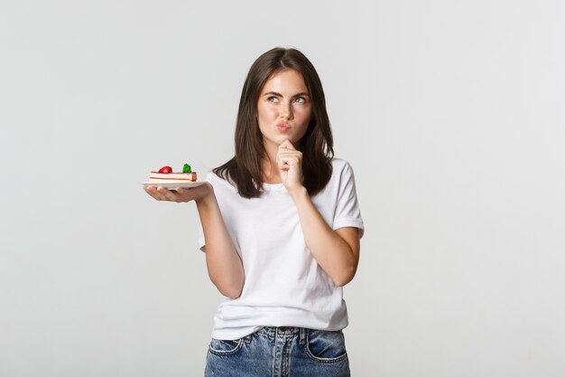 Rozważna uśmiechnięta ładna dziewczyna zamyślona trzymając kawałek ciasta, biały.