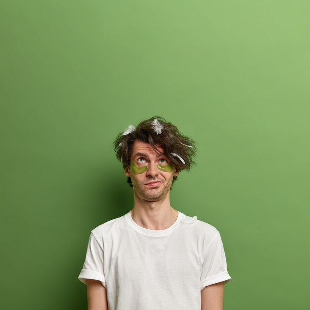 Bezpłatne zdjęcie rozważny mężczyzna skoncentrowany powyżej, czeka na ładny efekt po nałożeniu kolagenowych płatków pod oczy, ma rozczochrane włosy z piór, pozuje pod zieloną ścianą, skopiuj miejsce na twoją promocję