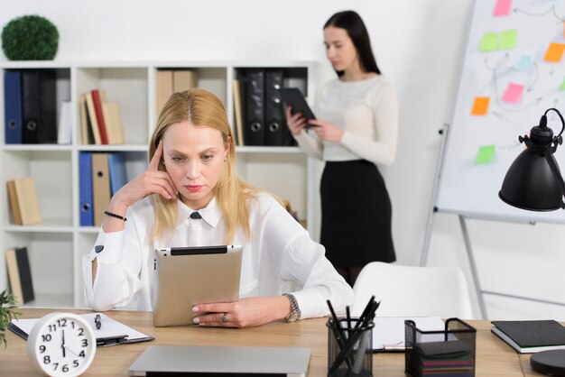 Rozważny młody bizneswoman patrzeje cyfrową pastylkę z jej kolega pozycją przy tłem w biurze