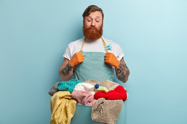 Bezpłatne zdjęcie rozważny wątpliwy niezdecydowany mężczyzna z gęstą rudą brodą, patrzy na pranie, nie umie prać, nie jest chętny do prania