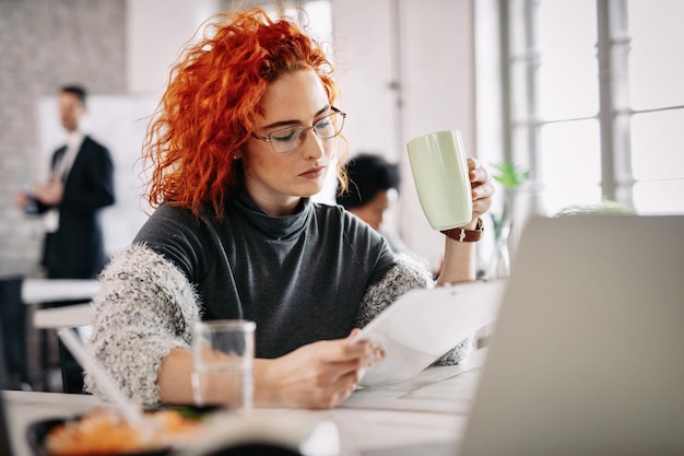 Ruda bizneswoman robi papierkową robotę pijąc kawę przy biurku w biurze W tle są ludzie