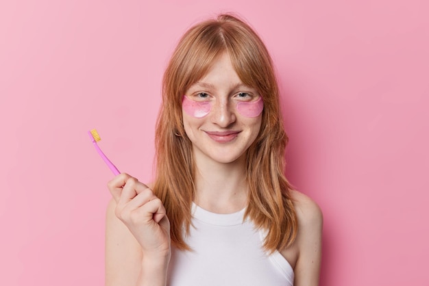 Bezpłatne zdjęcie ruda dziewczyna nakłada hydrożelowe plastry pod oczy trzyma szczoteczkę do zębów poddaje się zabiegom pielęgnacyjnym i higienicznym o poranku uśmiecha się przyjemnie na różowym tle dba o skórę i zęby