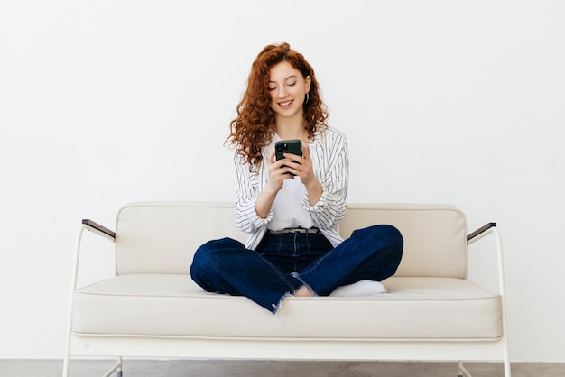 Ruda młoda kobieta leżąc na kanapie, korzystając z aplikacji online na nowoczesnym smartfonie, spędzając weekendowy dzień w domu