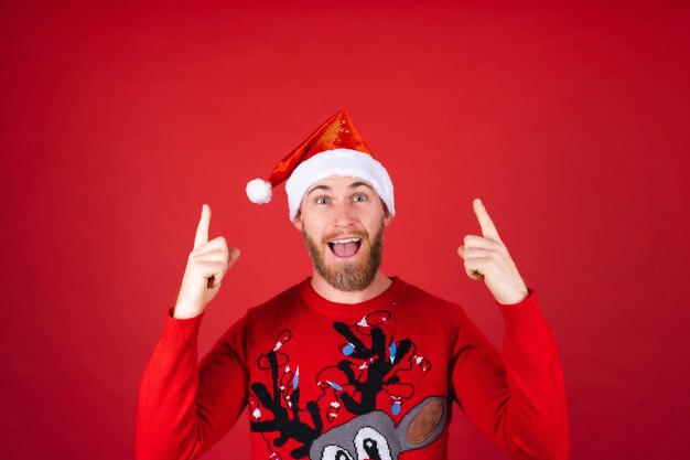 Rudobrody mężczyzna w czapce Mikołaja i świątecznym swetrze uśmiecha się radośnie wskazując palcem w górę