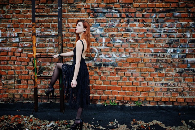 Rudowłosa dziewczyna punkowa nosi czarną sukienkę na dachu przy ceglanej ścianie z żelazną drabiną