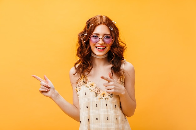 Rudowłosa kobieta w okularach przeciwsłonecznych wskazuje palcami w lewo. Kobieta w żółtej górze uśmiechając się na odosobnionym pomarańczowym tle.