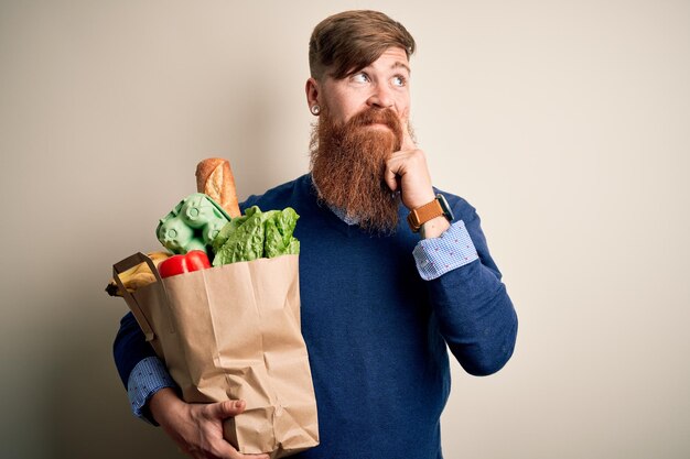Rudowłosy Irlandczyk z brodą trzymający świeże artykuły spożywcze z supermarketu na odosobnionym tle poważna twarz myśląca o pytaniu bardzo zdezorientowany pomysł