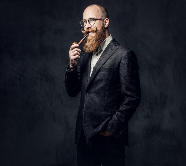 Rudy, brodaty mężczyzna ubrany w garnitur i okulary, palenie tradycyjnej fajki na ciemnoszarym tle.