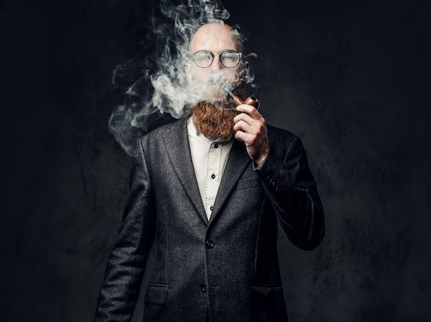 Bezpłatne zdjęcie rudy, brodaty mężczyzna ubrany w garnitur i okulary, palenie tradycyjnej fajki na ciemnoszarym tle.