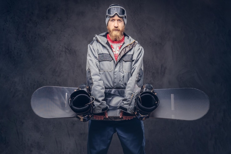 Bezpłatny plik Zdjęcie rudy brodaty snowboardzista w zimowej czapce i okularach ochronnych ubrany w snowboardowy płaszcz trzyma deskę snowboardową w studio. na białym tle na ciemnym tle z teksturą.