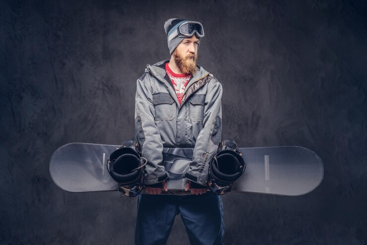 Bezpłatny plik Zdjęcie rudy brodaty snowboardzista w zimowej czapce i okularach ochronnych ubrany w snowboardowy płaszcz trzyma deskę snowboardową w studio. na białym tle na ciemnym tle z teksturą.