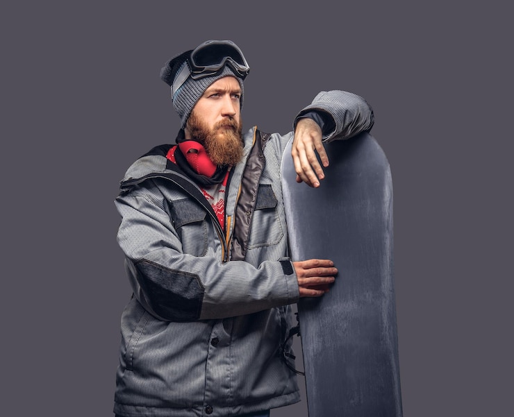 Bezpłatny plik Zdjęcie rudy brutalny snowboardzista z brodą w czapce zimowej i okularach ochronnych, ubrany w snowboardowy płaszcz, pozuje ze snowboardem w studio, odwracając wzrok. na białym tle na szarym tle.