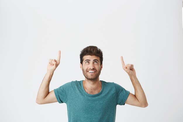 Salowy portret radosny brodaty hiszpański facet z zadowolonym wyrazem, ubrany w niebieski tshirt, śmiejący się i wskazujący do góry nogami na białej ścianie. Skopiuj miejsce