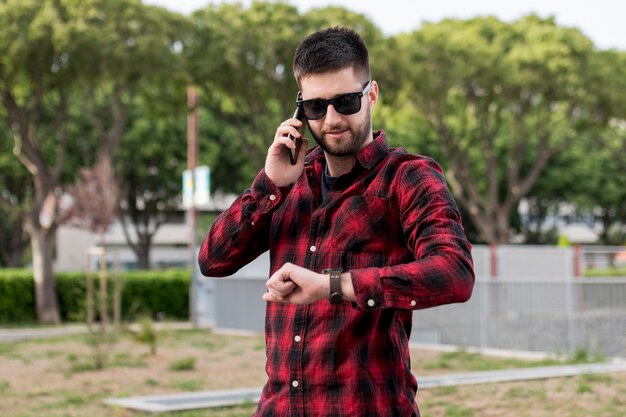 Samiec z okularami przeciwsłonecznymi trzyma smartphone blisko ucho i patrzeje zegarki