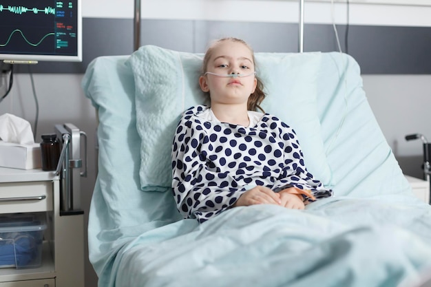 Bezpłatne zdjęcie samotna młoda blada chora pacjentka odpoczywa w łóżku kliniki pediatrycznej w pokoju na oddziale rekonwalescencji. chore dziecko samotnie siedzące w szpitalnym łóżku, noszące donosową rurkę tlenową do podtrzymywania życia.