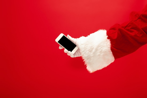 Santa Claus trzymając telefon komórkowy gotowy do Bożego Narodzenia na czerwonym tle studio. Sezon, zima, wakacje, uroczystość, koncepcja prezentu