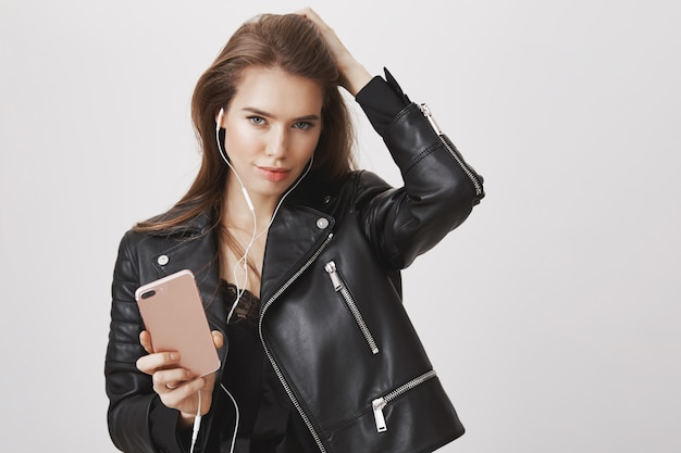 Sassy kobieta w skórzanej kurtce, nosi słuchawki i trzyma smartfon