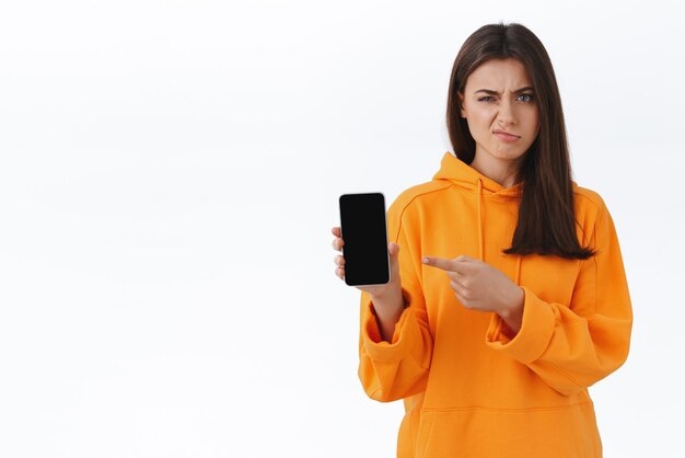 Sceptyczna brunetka narzeka na aplikację, która nie lubi złej obsługi firmy, którą pobrała trzymając telefon komórkowy i wskazując ekran smartfona z zawiedzionym grymasem na białym tle