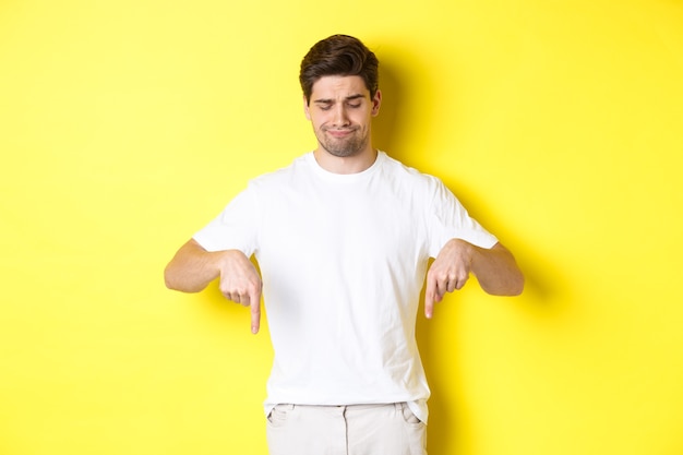 Sceptyczny młody człowiek w białej koszulce, wskazujący i spoglądający w dół zdenerwowany, dezaprobujący i nie lubiący produktu, stojący na żółtym tle.