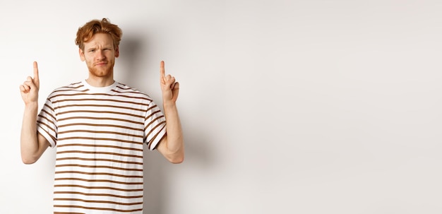 Bezpłatne zdjęcie sceptyczny przystojny rudy mężczyzna wskazujący palcami w górę, krzywiący się i patrzący niepewnie na stojącą kamerę
