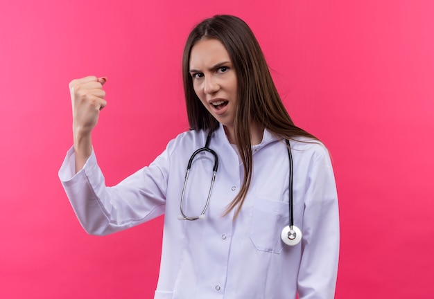 Ścisły młody lekarz dziewczyna ubrana w stetoskop medyczny fartuch robi silny gest na na białym tle różowym tle